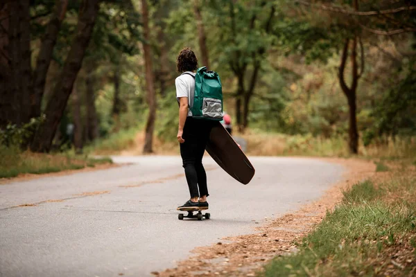 Visão traseira da jovem mulher ativa andando de skate com wakeboard em sua mão e mochila em seus ombros. — Fotografia de Stock