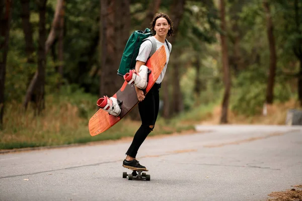 Actief meisje rijden skateboard met wakeboard in haar hand en rugzak op haar schouders. — Stockfoto