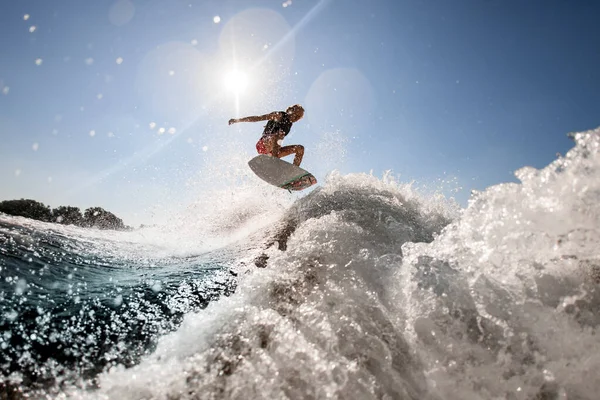 Man professionally rides and bounces on surf style wakeboard over splashing wave against blue sky — Stock Photo, Image