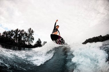 Siyah mayo giymiş genç enerjik bir kadın sörf tahtasında dalgaların üzerinde zıplıyor..