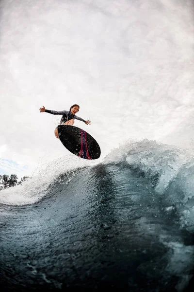 Mujer activa despierta surfista en traje de neopreno negro salta efectivamente en la tabla de surf en la ola —  Fotos de Stock