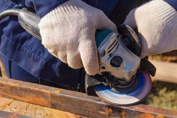 A worker in overalls is grinding a weld seam on a steel square profile pipe with an angle grinder with a flap disc.