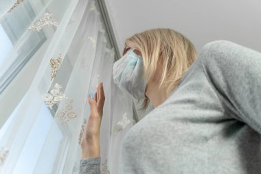 Evde izole edilmiş, maske takmış, camdan dışarı bakan üzgün sarışın kız.