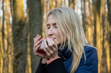 Genç sarışın bir kız gün batımında bahar ormanında termos bardağından çay içer. Tema yolculuğu. Yürüyüş sırasında çay içen kız.