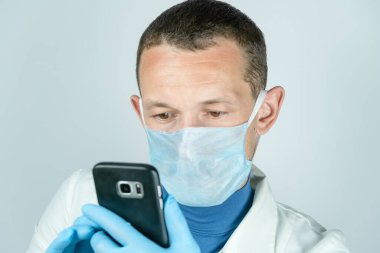 Siyah bir cep telefonu tutan, tıbbi önlüklü, maskeli ve eldivenli bir erkek doktorun portresi.