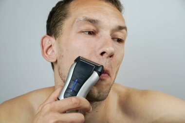 Açık arkaplanda elektrikli jileti olan koyu renk saçlı güzel bir adamın portresi. Koyu renk saçlı adam yakın plan elektrikli tıraş makinesiyle tıraş oluyor..