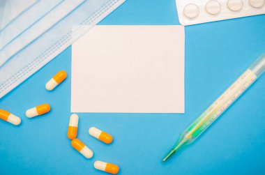 Tıbbi maske, turuncu ve beyaz kapsüller, termometre, mavi arka planda haplar ve fotokopi alanı olan beyaz bir sayfa. İlk yardım, hastalık sırasında bakım..