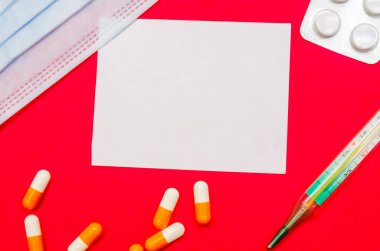 Tıbbi maske, turuncu ve beyaz kapsüller, termometre, kırmızı arka planda haplar ve fotokopi alanı olan beyaz bir sayfa. İlk yardım, hastalık sırasında bakım..