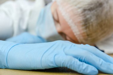 Tıbbi maskeli, tıbbi şapkalı ve mavi eldivenli yorgun kadın doktor yakın planda bir masada uyukluyor. Tıp kavramı