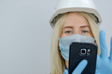 Tıbbi maskeli ve beyaz miğferli genç sarışın kadın siyah bir cep telefonuna bakar.