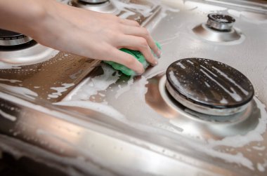 Kadının eli yeşil köpüklü süngerle gri paslanmaz çelik bir gaz sobasını yıkıyor. Ev temizliği