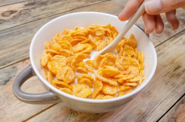 Beyaz bir kaşıkla kızın eli mısır gevreğini tahta bir arka planda sütle karıştırıyor.