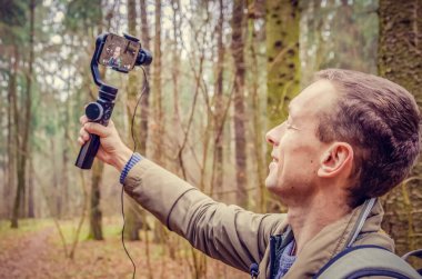 Erkek blogcu kendini ormanda cep telefonu kamerasıyla çekiyor ve kameraya gülümsüyor ve el sallıyor. Arkadan çekilen fotoğraf. blog yazma, internet, bilgi kavramı