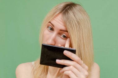 Genç bir kadın aynaya bakar ve sivilceleri ve morlukları düzeltmek için kapatıcı kullanır. yeşil bir arkaplanda