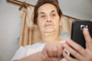 Siyah saçlı yaşlı bir kadın önünde siyah bir cep telefonu tutuyor, ona bakıyor ve parmağını ekrana vuruyor.