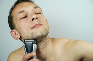 Açık arkaplanda elektrikli jileti olan koyu renk saçlı güzel bir adamın portresi. Koyu renk saçlı adam yakın plan elektrikli tıraş makinesiyle tıraş oluyor..