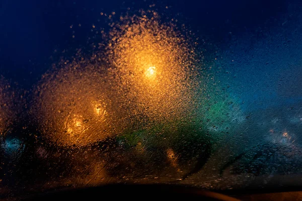 街の通りの光が透明ガラスを通して見えます水からの霧が濃い青の夜に落ち虹色をしていて銀河空間の多くの星のように見えます — ストック写真