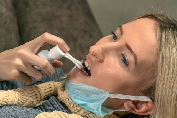 young blonde woman with a scarf around her neck and a medical mask pulled down on her chin is lying on the sofa and stabbing her throat with a spray