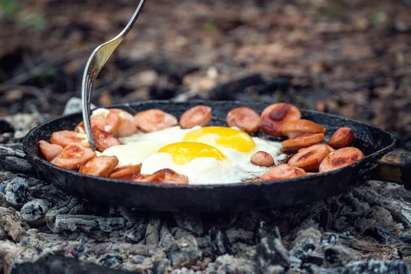 Girl\'s hand with a fork turns over pieces of fried sausage and eggs in a pan on the coals close up. Breakfast in nature, picnic, camping food.