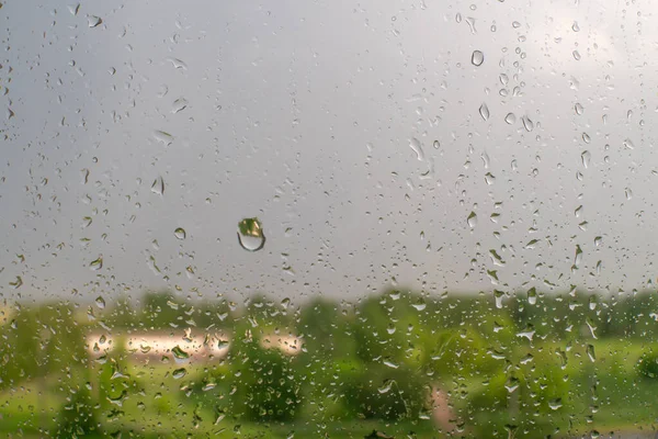 Raindrops on a window with green trees and a cloudy sky in the background on a summer day