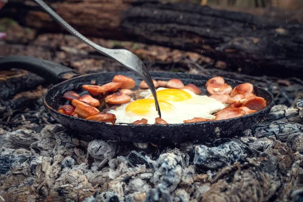 Girl\'s hand with a fork turns over pieces of fried sausage and eggs in a pan on the coals. Breakfast in nature, picnic, camping food.