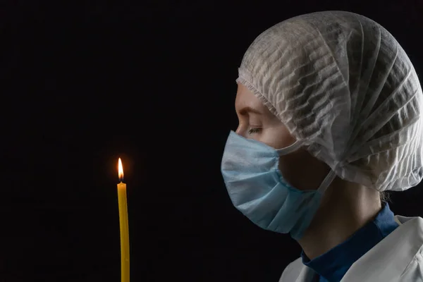 young woman stands sideways in a medical mask, medical cap and white coat holding a burning candle in front of her close-up on a black background. Concept of faith in a bright future.