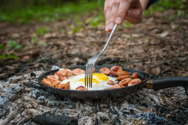 Girl\'s hand with a fork turns over pieces of fried sausage and eggs in a pan on the coals. Breakfast in nature, picnic, camping food.