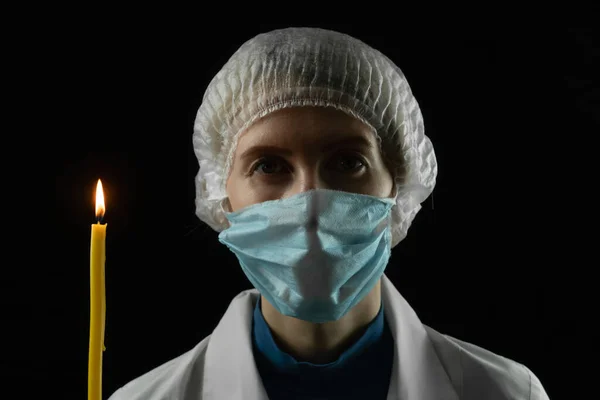 Young woman in a medical mask, medical cap and white coat holds a burning candle in front of her on a black background. Concept of last hope, faith in a bright future.