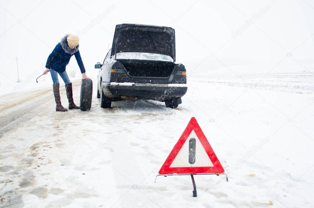 woman changing the wheel of a black car with an open trunk in winter snow with the alert sign on a snowy road
