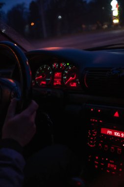 Sürücünün elleri arabanın direksiyonunda, gece araba sürerken. Kırmızı ışıklı araba konsoluna odaklan. Dikey fotoğraf