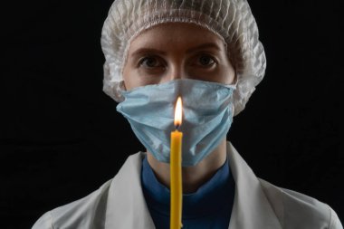 Tıbbi maskeli, tıbbi şapkalı ve beyaz önlüklü genç bir kadın siyah arka planda yanan bir mum tutuyor. Son umut kavramı, parlak bir geleceğe olan inanç. Kadın doktora odaklan.