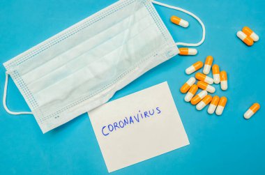 Tıbbi maske, mavi arka planda turuncu-beyaz kapsüller ve beyaz bir sayfada Coronavirus yazılıydı. İlk yardım, hastalık sırasında bakım..