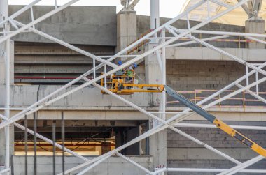 Ressam, özel elbiseli sıva ustası, mavi gökyüzüne karşı güneşli bir günde beşikte beyaz boya silindiriyle binanın ön cephesini boyuyor..
