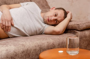 Bir adam kanepeye uzanıp bir bardak su ve hapa bakar. Hastalık konsepti