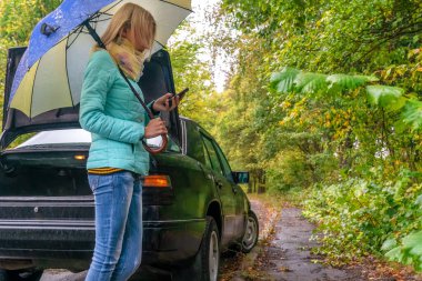 İnce, sarışın bir kız yağmur altında kırık bir arabanın yanında duruyor ve yardım çağırmak için bir cep telefonuna bakıyor.