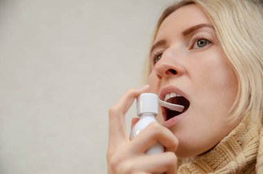 Boğazında fular olan sarışın bir kız boğazı ağrıdığı için ağzına sprey sıkar. Sağlık ve tıp kavramı. Boşluğu kopyala.