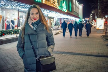 Genç, güzel, kahverengi saçlı, uzun saçlı, dudaklarında kırmızı ruj olan, gülümseyen ve geceleri şehirde yürürken kameraya bakan bir kız.