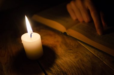 Açık bir kitap, bir kitap üzerinde parmak, ve karanlık bir masada yanan bir mum.