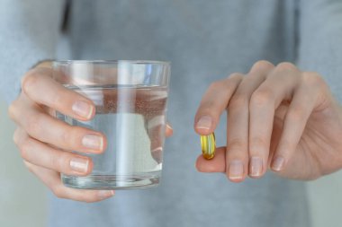 Gri ceketli kadın elinde bir omega-3 balık yağı kapsülü ve bir bardak su tutuyor. Biyolojik olarak aktif katkı maddesi. E vitamini, beriberi 'ye karşı altın bir tablet..