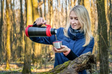 Gezgin sarışın kız bahar ormanındaki bir termostan çay dolduruyor. Tema yolculuğu. Bir kadın termostan bir bardağa sıcak içecek dolduruyor..
