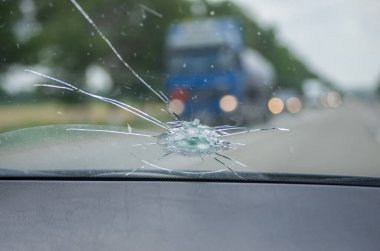 Uçan taştan arabanın kırık ön camı. Camdaki delik, kırıntılar ve molozlar, şeritler halinde çatlaklar. Cam bulutlarla gökyüzünü yansıtıyor..