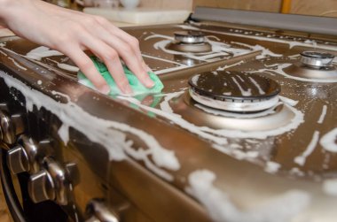 Kadının eli yeşil köpüklü süngerle gri paslanmaz çelik bir gaz sobasını yıkıyor. Ev temizliği