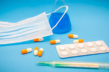 Tıbbi maske, turuncu ve beyaz kapsüller, mavi sıvılı matara, termometre, mavi arka planda tabletler. Tıp ve bilim kavramı. Arkaplan