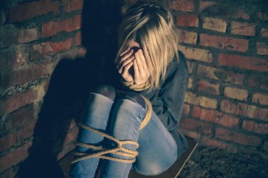 blonde girl with bound hands and feet is sitting in the basement, covering her face with her hands and crying. Concept of violence, kidnapping clipart