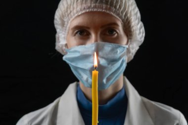 Tıbbi maskeli, tıbbi şapkalı ve beyaz önlüklü genç bir kadın siyah arka planda yanan bir mum tutuyor. Son umut kavramı, parlak bir geleceğe olan inanç.