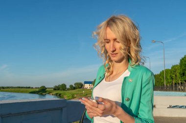 Kıvırcık saçlı sarışın kız elinde cep telefonuyla nehrin kenarında duruyor.