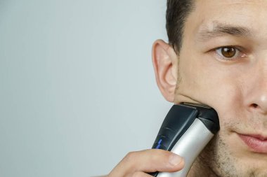 Yanağında elektrikli tıraş bıçağı olan yakışıklı, koyu saçlı bir adamın yüzünün yarısı, kopyalama alanı. Elektrikli jiletle tıraş olmak..