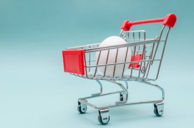 Mavi arka planda alışveriş arabasında beyaz tavuk yumurtası. Tüketici konsepti, minimalist online alışveriş. Boşluğu kopyala