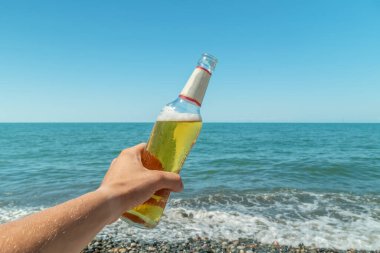 Bir adamın eli, güneşli bir günde deniz ve mavi gökyüzüne karşı bir şişe bira tutar. Dinlenme konsepti.