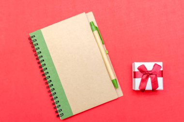 Kırmızı arka planda kalem ve hediye olan not defteri. Düz konum, üst manzara. Tatil konsepti, Sevgililer Günü, Noel, Yeni Yıl